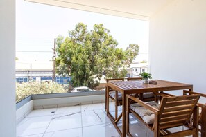 A balcony with a wooden table and chairs, a potted plant, and a view of a tree and buildings. - Platinum Breeze - 1-BR Apt. in Larnaca (Larnaka)