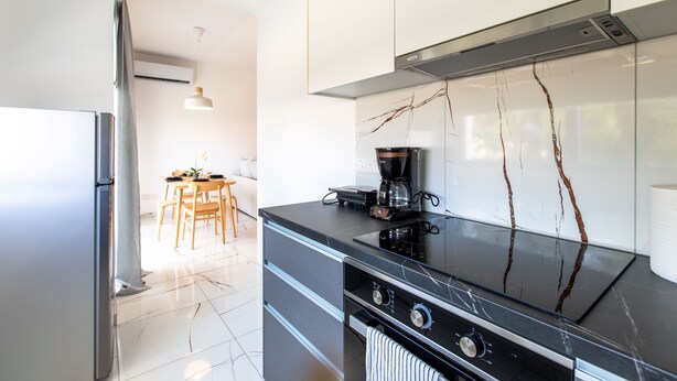 A modern kitchen with a built-in oven, a coffee maker, and a dining area with a table and chairs. - Platinum Breeze - 1-BR Apt. in Larnaca (Larnaka)