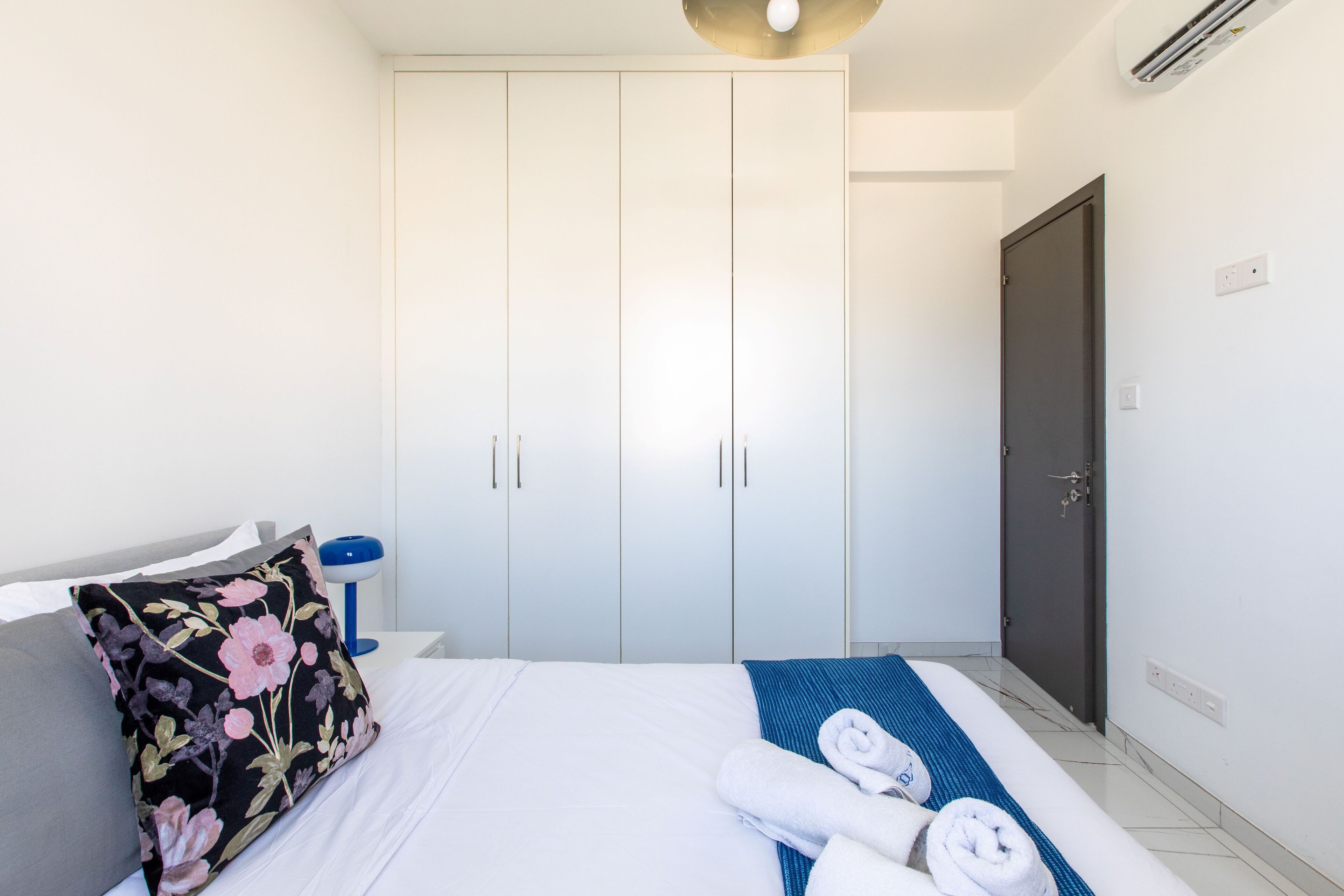 A hotel room with a bed, a wardrobe, a door, and a bedside table with a lamp.
