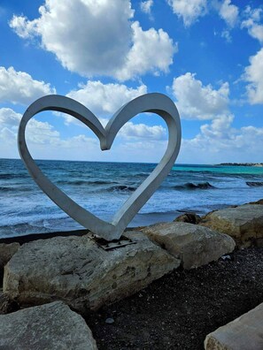 A heart-shaped metal sculpture on a rocky shoreline with the ocean and a clear sky in the background. - Studio in the heart of Paphos (Pafos)