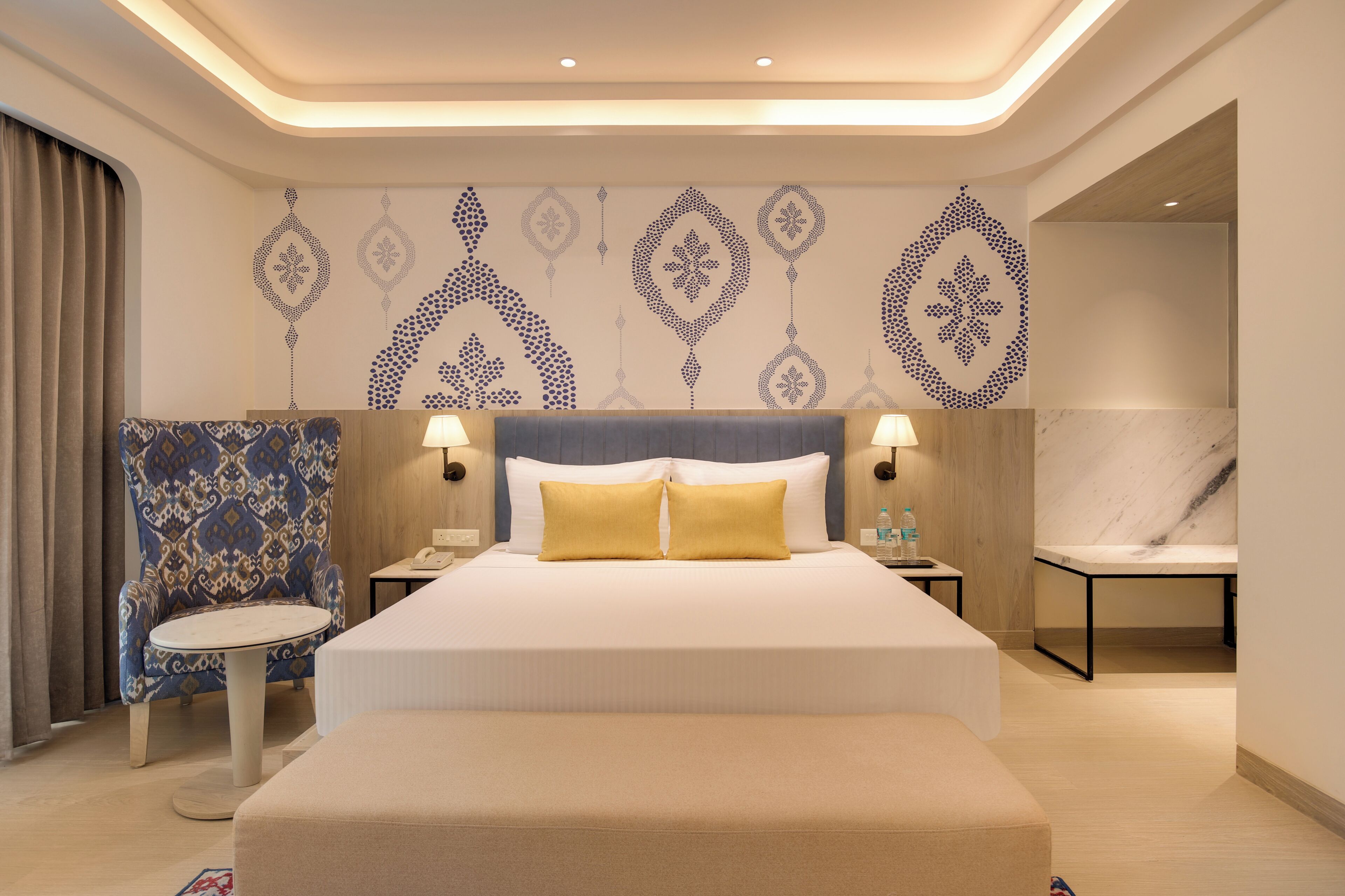 Una habitación de hotel con una cama grande, dos mesitas de noche con lámparas, una silla y un papel tapiz estampado.