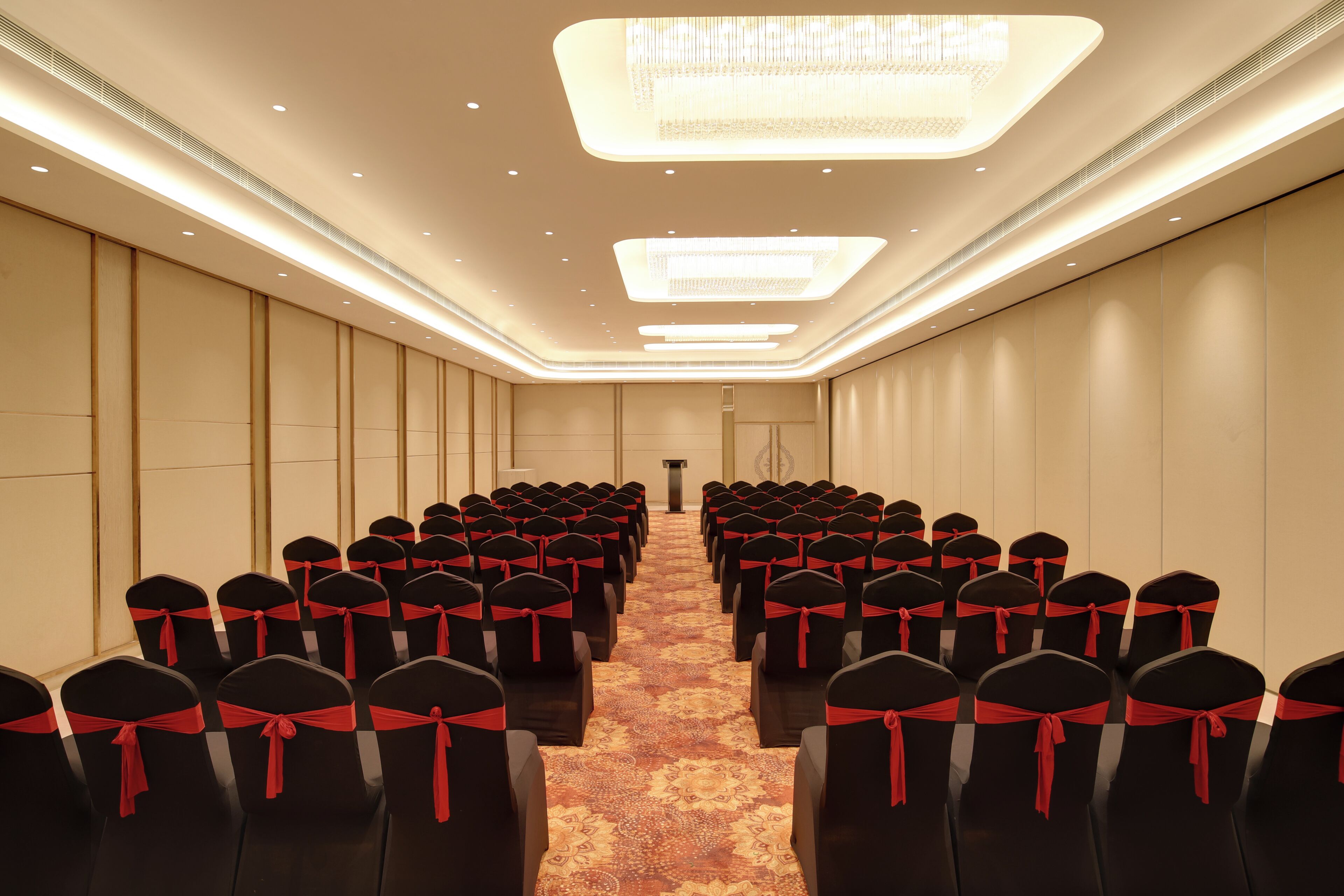 Un salón de conferencias con filas de sillas, cada una adornada con una cinta roja. El salón tiene una alfombra estampada y una lámpara de araña moderna.