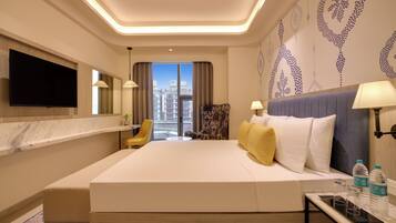 A hotel room with a large bed, a TV, a desk with a lamp, a chair, and a view of buildings through the window.