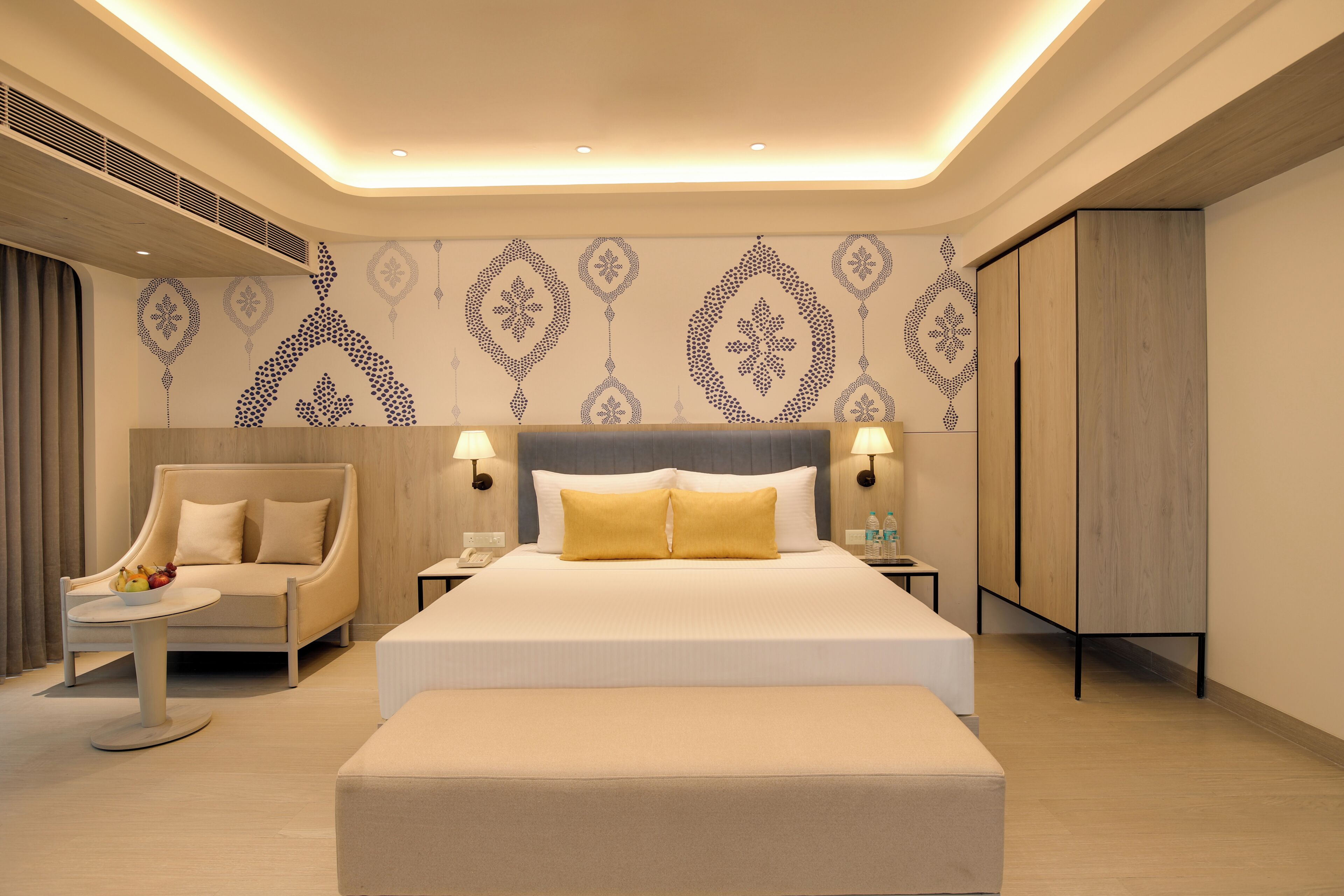 Una habitación de hotel moderna con una cama grande, lámparas de noche, una zona de estar con sofá y sillón, y patrones decorativos en la pared.