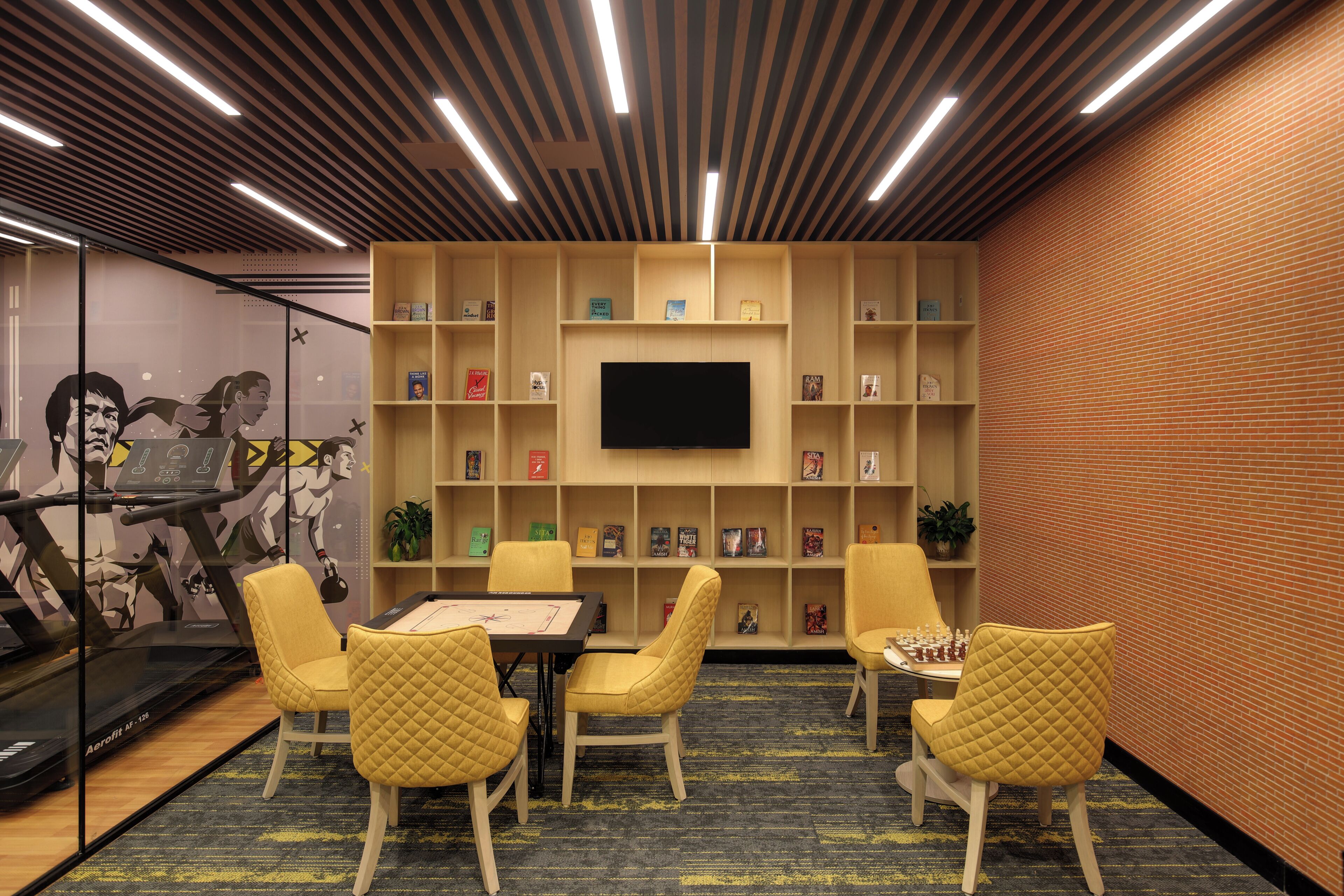 A room with a large flat-screen TV mounted on a brick wall, surrounded by bookshelves. There are four yellow chairs and a table with a chessboard.
