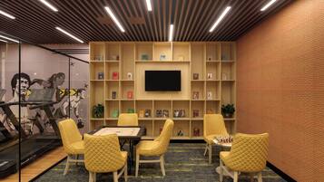 A room with a large flat-screen TV mounted on a brick wall, surrounded by bookshelves. There are four yellow chairs and a table with a chessboard.