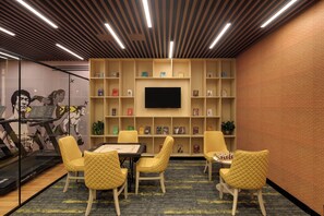 A room with a large flat-screen TV mounted on a brick wall, surrounded by bookshelves. There are four yellow chairs and a table with a chessboard.