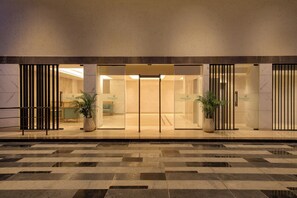 Entrada de un hotel con puerta de vidrio, plantas en macetas y un suelo con diseño a cuadros.