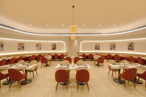 A spacious dining area with round tables and chairs, recessed lighting, and wall art. -  Lemon Tree Hotel, Bharuch (Bharuch)