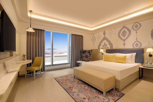 A modern hotel room with a large bed, a desk, a chair, and a view of the sky.