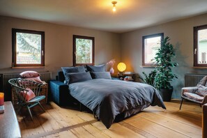 A bedroom with a bed, two windows, a radiator, a chair, a bedside table with a lamp, and a potted plant. - Hofzehn – for Groups & Families (Trautskirchen)