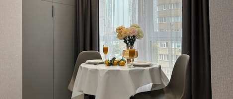 A dining area with a table set for two, a vase with flowers, and a city view through sheer curtains.