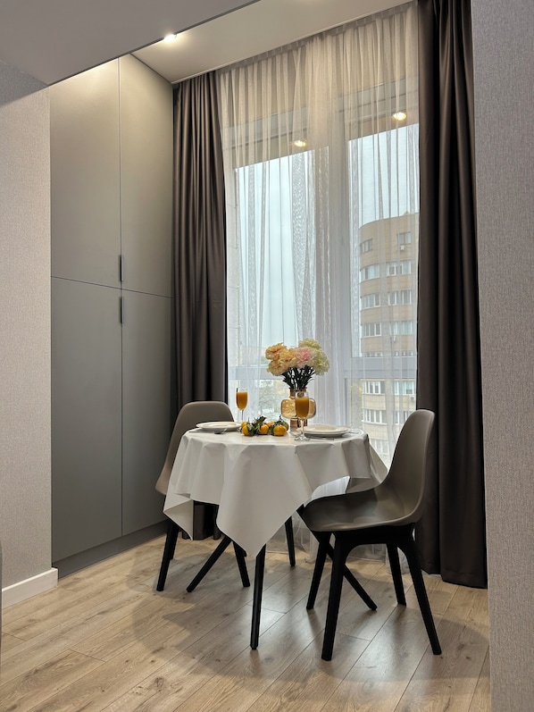 A dining area with a table set for two, a vase with flowers, and a city view through sheer curtains. - Comfort city Residence near to MEGA (Almaty)