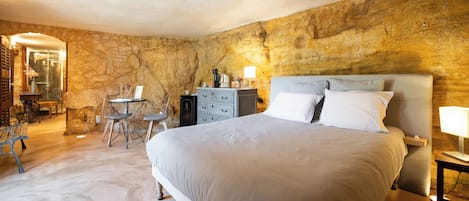A bedroom with a stone wall, a bed with white bedding, a small table with a lamp, and a chair.