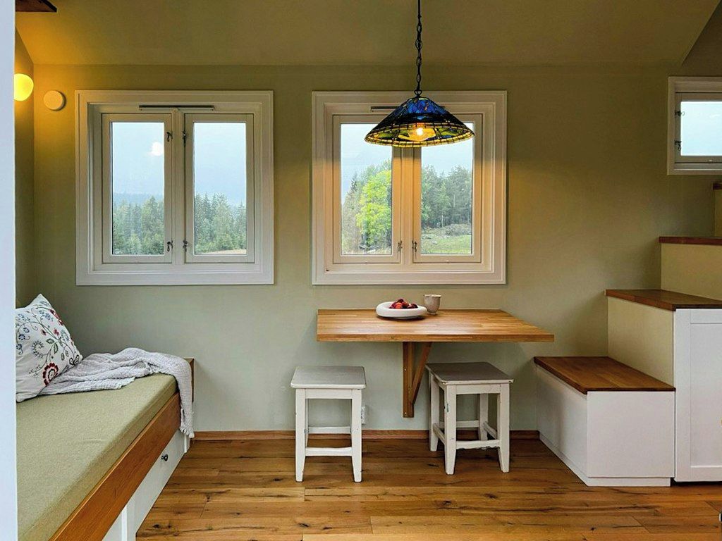 A compact room with a bed, a small table with stools, a window, and a hanging light fixture.