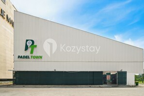 A large industrial building with a flat facade, featuring the 'Kozystay' and 'Padel Town' logos. - Ravora by Kozystay - Sungkono (Surabaya)