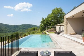 Ein rechteckiger Pool mit klar blauem Wasser, umgeben von einer gepflasterten Liegefläche mit Liegestühlen und einem Tisch.