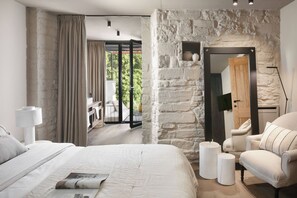 Ein Schlafzimmer mit Steinswand, einem Bett mit Zeitschrift, einem Sessel und Blick ins Freie.