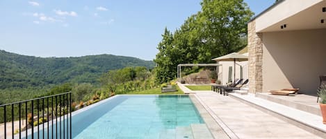 Una piscina infinita rectangular con agua azul cristalina, rodeada por una terraza de piedra y barandillas de metal negro.
