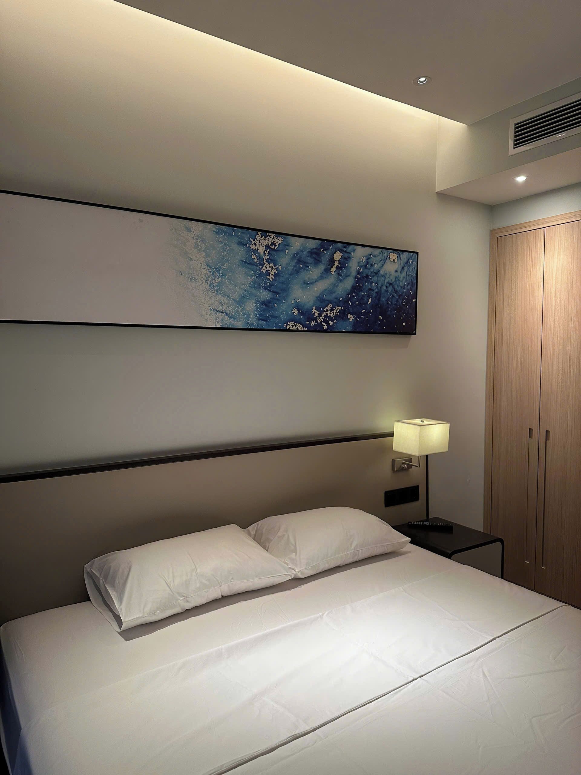 Une chambre d’hôtel avec un lit, une table de chevet avec une lampe, une armoire et un tableau accroché au mur.