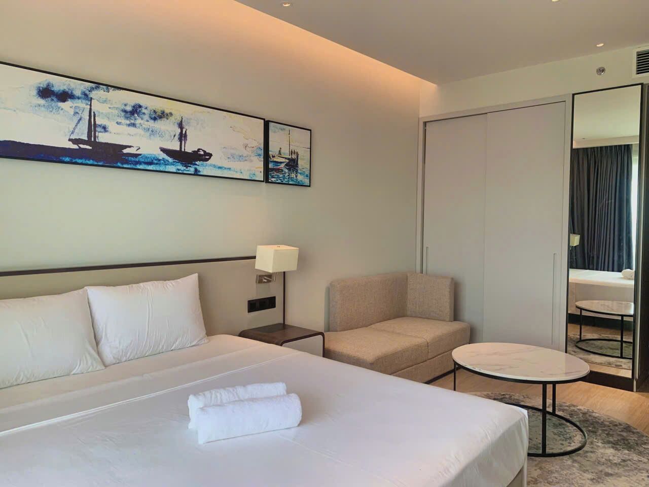 A hotel room with a large bed, a sofa, a small round table, a lamp, and a painting on the wall.