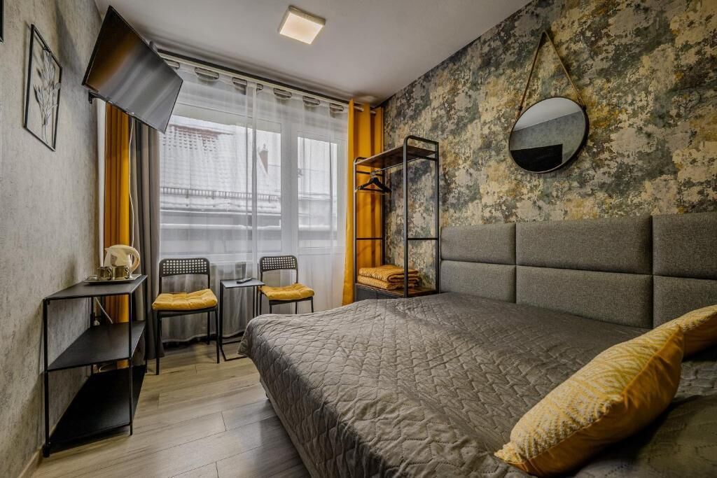 Um quarto com uma cama, mesa de cabeceira, uma cadeira, uma mesinha e uma janela com cortinas.