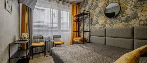 Um quarto com uma cama, mesa de cabeceira, uma cadeira, uma mesinha e uma janela com cortinas.