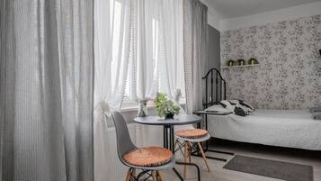 A bedroom with a bed, a small table, two chairs, a window with sheer curtains, and a wall with floral wallpaper.