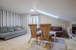 A loft-style living room with a sloped ceiling, a grey sofa, a brown sofa, wicker chairs, a small white table, and a television.