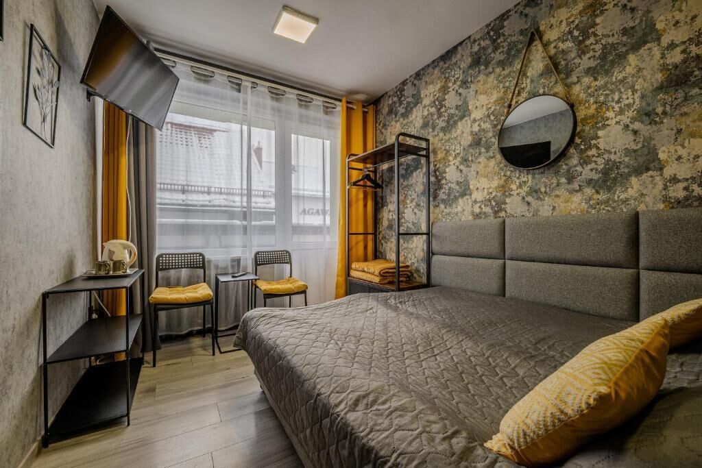 Un dormitorio con cama, mesita de noche, silla, pequeña mesa, espejo y ventana con cortinas.