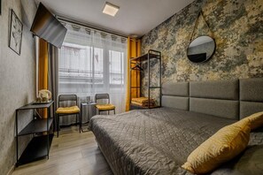 A bedroom with a bed, a bedside table, a chair, a small table, a mirror, and a window with curtains.