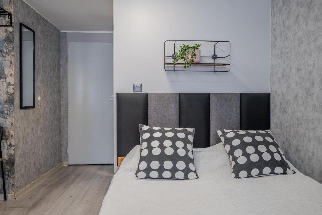 Un dormitorio con cama, dos almohadas, una mesita de noche, un estante en la pared con una planta y una puerta.