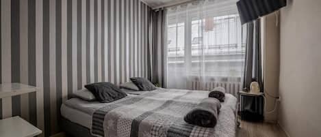 A modern bedroom with a bed, a bedside table, a flat-screen TV, and a window with curtains.