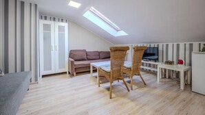 A room with a sloped ceiling, a sofa, a dining table with chairs, a television, and a kitchenette area.