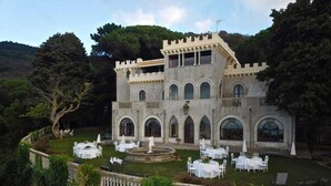 Un edificio con aspecto de castillo, con varios balcones y ventanas de arco, rodeado de un jardín cuidado con mesas y sillas dispuestas para comer al aire libre.