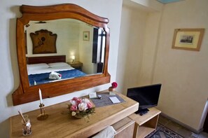Habitación de hotel con un espejo grande, una cama con ropa de cama blanca, una mesita de noche con lámpara, un jarrón de flores y un televisor.