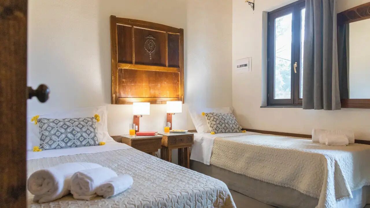A hotel room with two beds, a wooden headboard, a window with curtains, and a bedside table with a lamp.