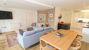 A modern living room with a sofa, a dining table with chairs, a kitchen area, and a flat-screen TV mounted on the wall. - 11 Fountain Court (Falmouth)