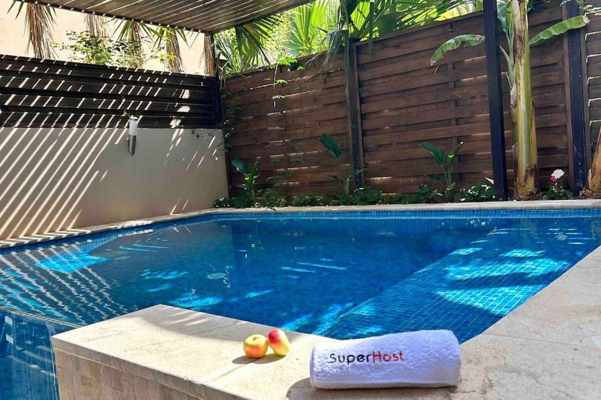 A swimming pool with a wooden fence, a towel labeled 'SuperHost,' and two apples on the edge.