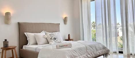 A modern bedroom with a large bed, two bedside tables, and a view of the outdoors through sheer curtains.