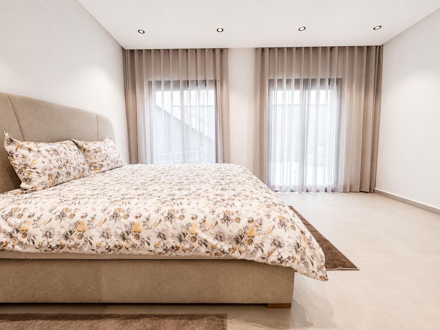 Ein modernes Schlafzimmer mit einem großen Bett, zwei Fenstern mit durchsichtigen Vorhängen und Deckeneinbaustrahlern.