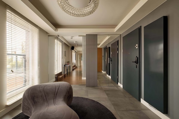 A hotel corridor with a modern design, featuring a seating area with a grey upholstered chair and a dark rug, leading to a series of doors and a staircase. - 154 sqm | Chmielna 63 Penthouse | 3 Bedrooms | AC (Gdańsk)