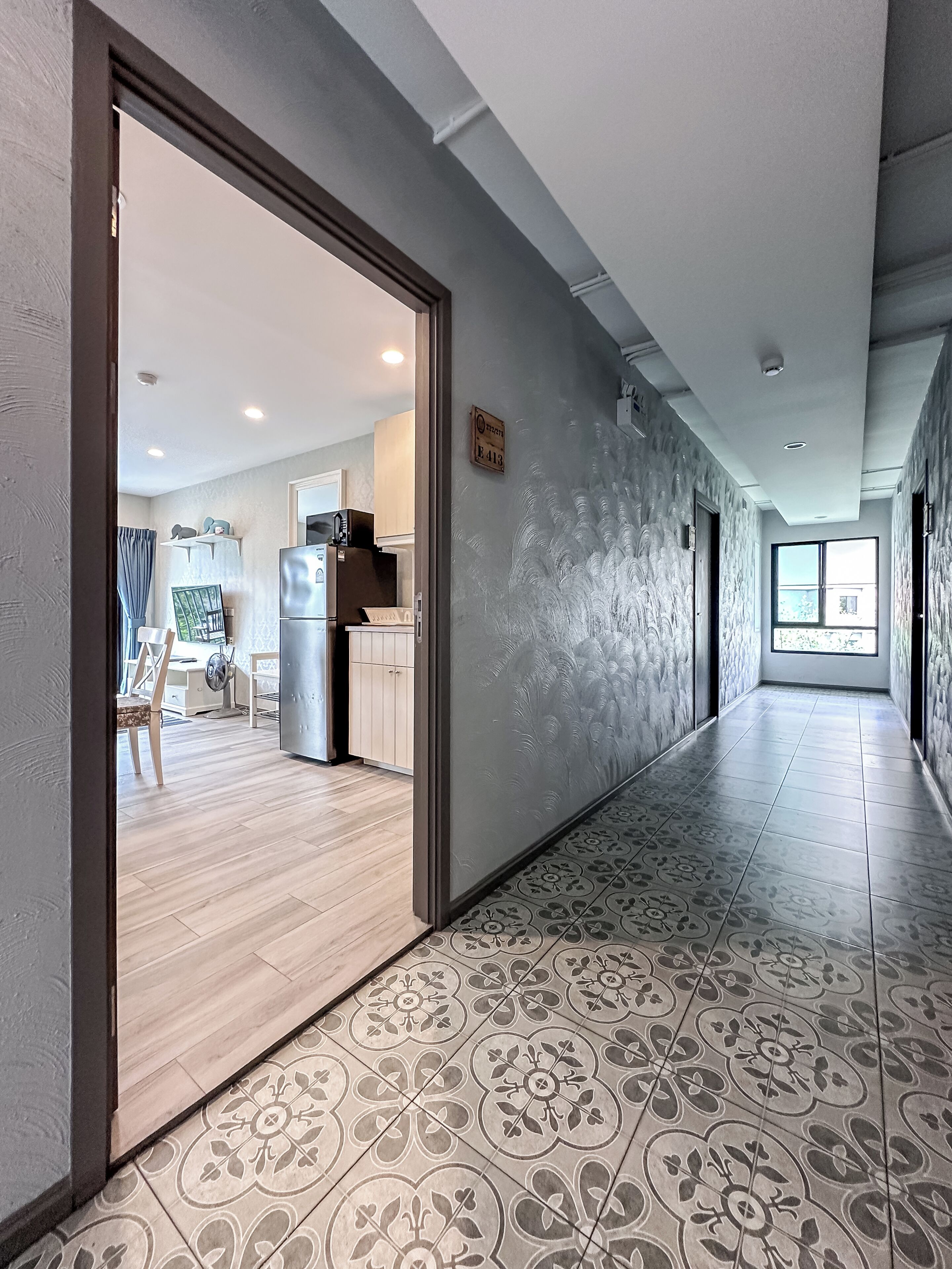 Un pasillo con piso estampado, una zona de cocina con armarios y electrodomésticos, y una puerta que da a otra habitación.