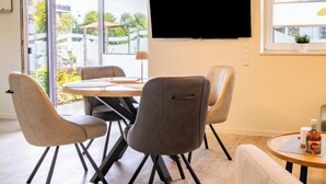 A dining area with a round table, four chairs, a flat-screen TV mounted on the wall, and a window with a view of the outside. - Coastal Lodge 33/1 - Just a step from the sea. Sauna. Patio. Garden. (Tossens)