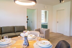 Ein modernes Wohnzimmer mit Essbereich, ausgestattet mit einer Couch, einem Sessel, einem Esstisch gedeckt mit Tellern, Gläsern und einer Vase mit Blumen.