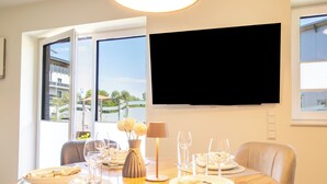 A dining area with a table set for four, a flat-screen TV mounted on the wall, and large windows offering a view of buildings and trees. - Coastal Lodge 33/1 - Just a step from the sea. Sauna. Patio. Garden. (Tossens)