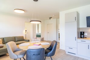 A modern living room with a sofa, two armchairs, a coffee table, and a dining table. There is a kitchen area with cabinets and a refrigerator. - Coastal Lodge 33/1 - Just a step from the sea. Sauna. Patio. Garden. (Tossens)