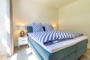 A neatly made bed with striped bedding, a bedside table with a lamp, and a window with curtains. - Coastal Lodge 33/1 - Just a step from the sea. Sauna. Patio. Garden. (Tossens)