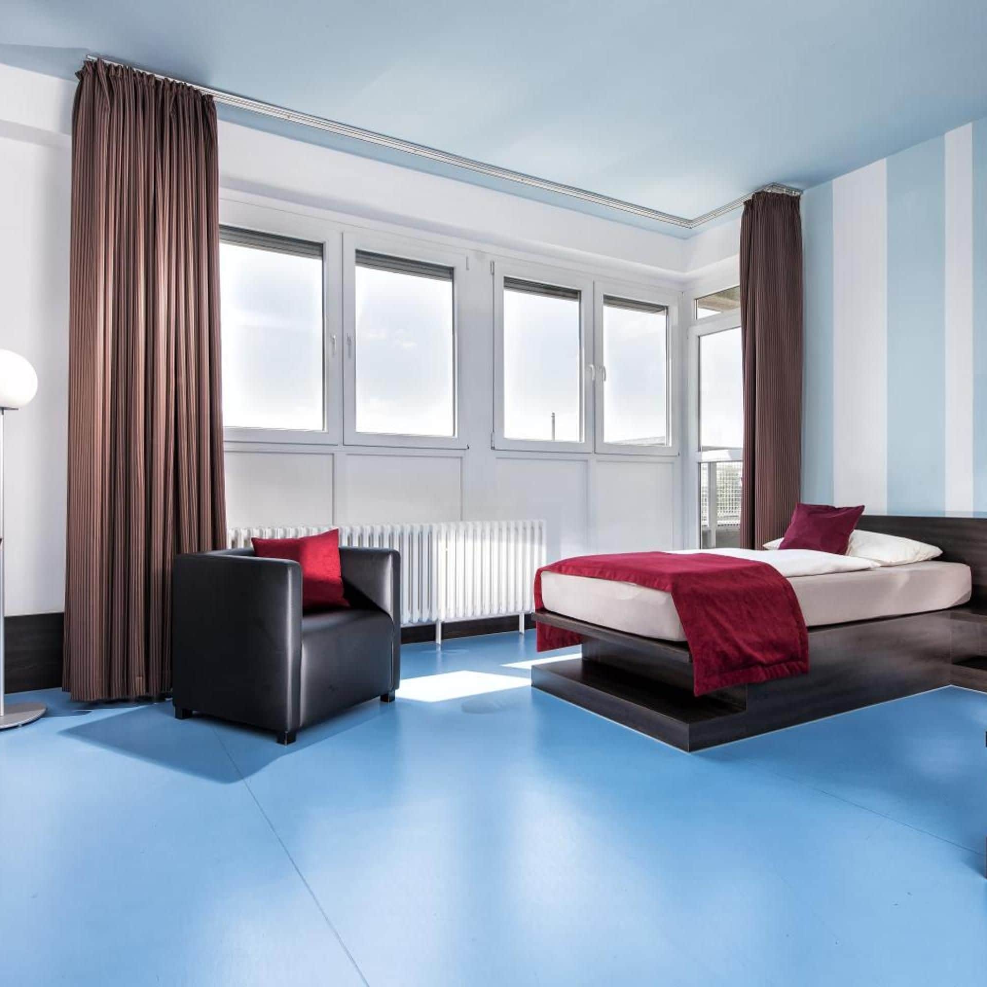 Ein modernes Hotelzimmer mit Bett, einer Sitzecke, einem Sessel und großen Fenstern.