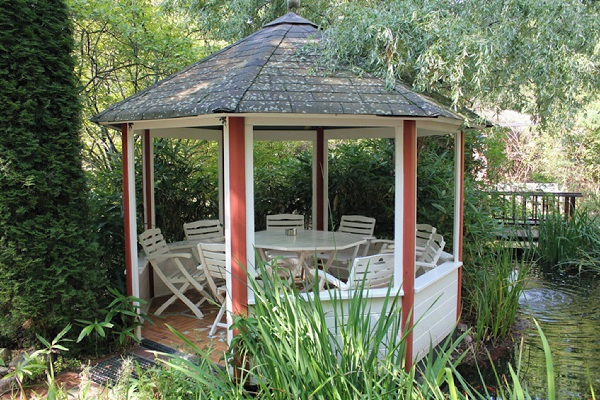 Ein Gazebo mit einem Ziegeldach, umgeben von Grünpflanzen und einem Teich.